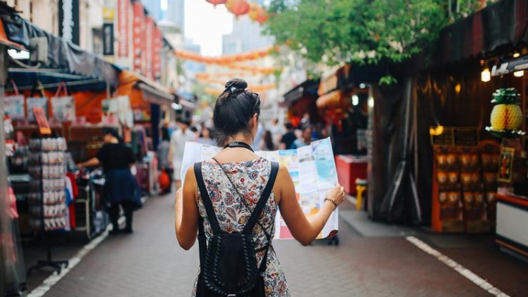 Rear view image of a young brunette woman. She is enjoying the walk and exploring the city, wearing a casual but fashionable dress, sightseeing and shopping on the Singapore street market. She is holding a large city map, checking out where to go next.