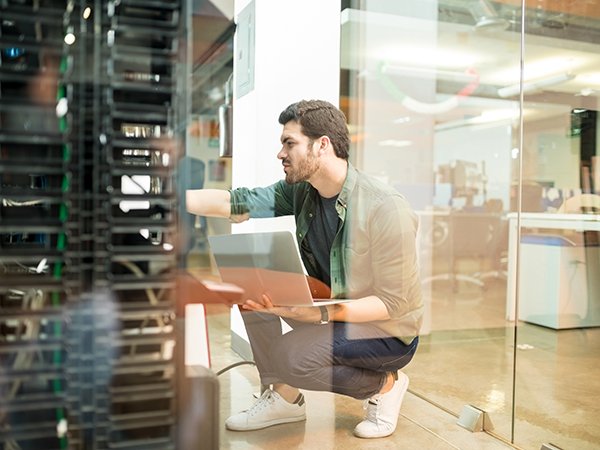 Network administrator with notebook computer sitting in data center room and working with networking device on rack cabinet